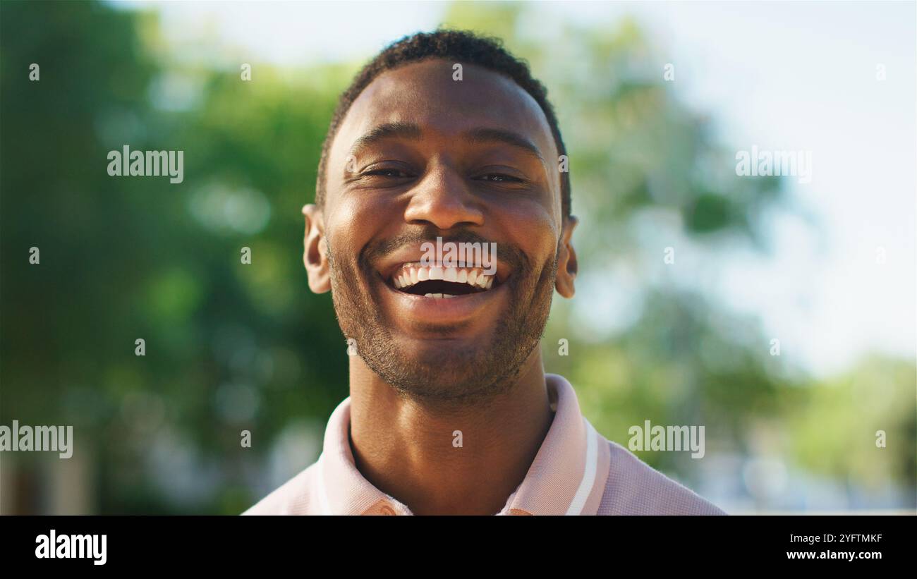 Un giovane allegro si erge all'aperto, sorridendo radioso mentre la luce del sole filtra tra gli alberi sullo sfondo. Trasuda positività e warm Foto Stock