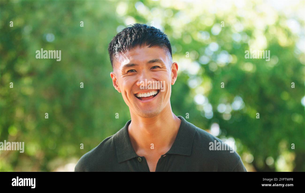 Un uomo allegro si trova all'aperto, sorridendo con un ampio sorriso. Indossa una camicia verde e ha i capelli corti, con la luce del sole che filtra attraverso le foglie Foto Stock