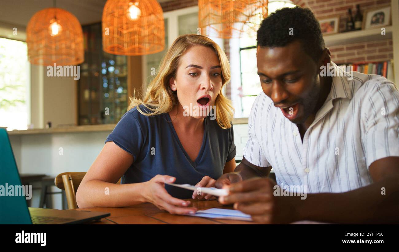 Una coppia si siede su un tavolo di legno, esprimendo gioia e sorpresa mentre guarda il loro dispositivo mobile. La camera, ben illuminata, è arredata in modo elegante e caldo Foto Stock
