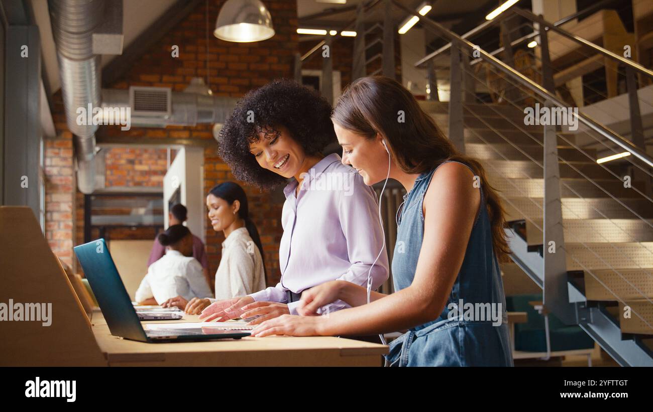 Donna d'affari che incontra una collega donna che lavora su un computer portatile e ascolta musica tramite auricolari Foto Stock