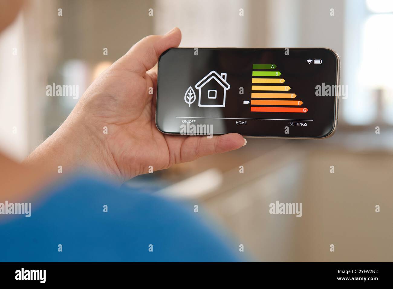 Una persona ha in mano uno smartphone con grafica colorata che rappresenta i livelli di efficienza energetica domestica. L'ambiente è uno spazio abitativo contemporaneo, Foto Stock