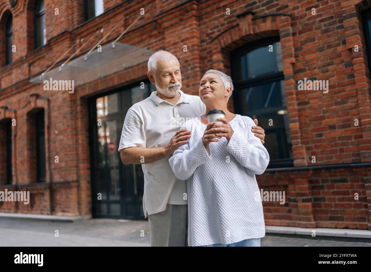 Affascinante uomo e donna anziana in piedi su strada asfaltata urbana, tenendo tazze di caffè e amando guardarsi l'un l'altro. Felice coppia senior che si diverte Foto Stock