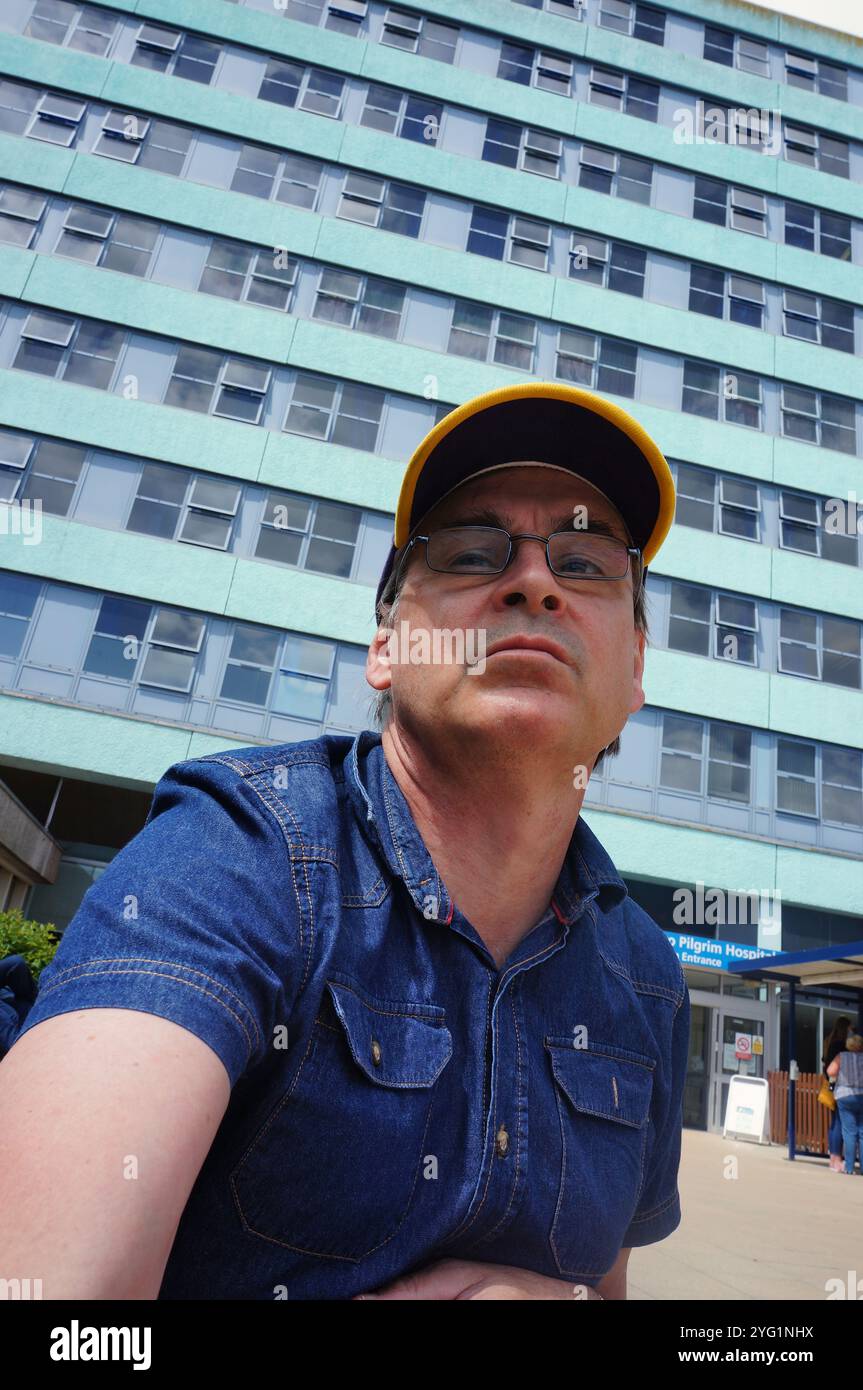 un uomo maturo che indossa un berretto da baseball e occhiali in piedi davanti a un edificio a più piani. Foto Stock