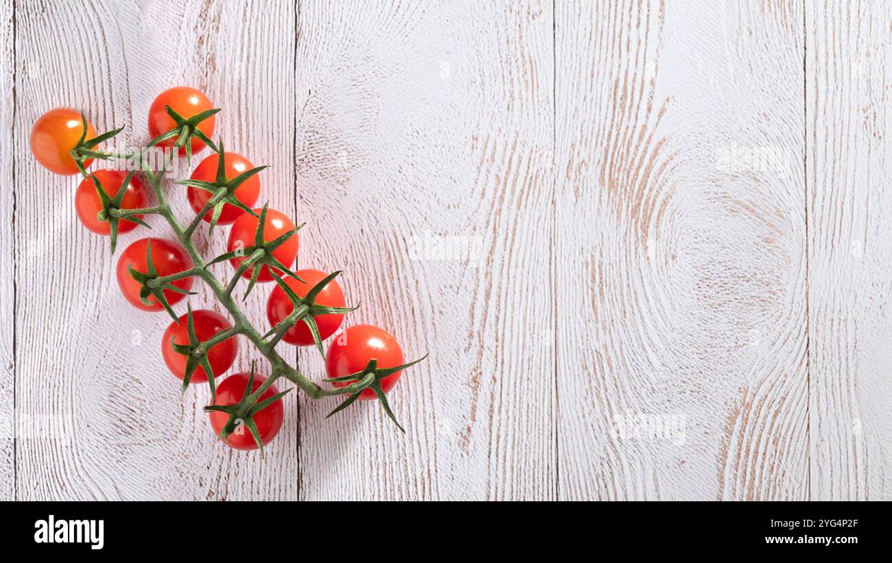 Il gruppo di pomodori ciliegini sulla vite poggia su una superficie in legno bianco testurizzato, evidenziando il contrasto tra i pomodori rossi accesi e. Foto Stock