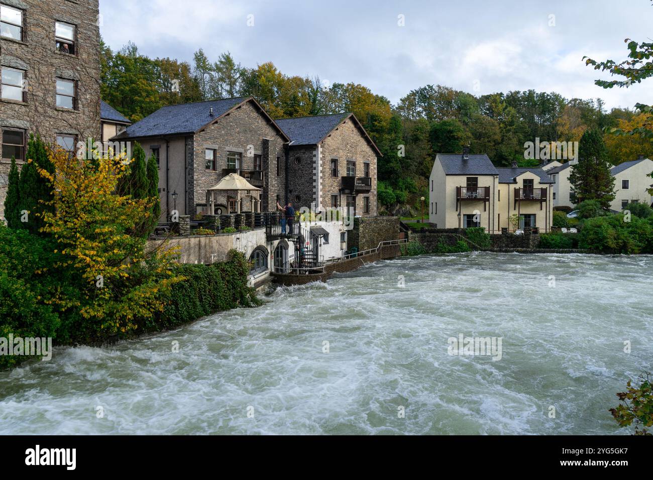 Case moderne a Backbarrow, Cumbria, Regno Unito, circondate da una vivace vegetazione autunnale. Il fiume Leven, che è in piena, scorre attraverso il villaggio Foto Stock