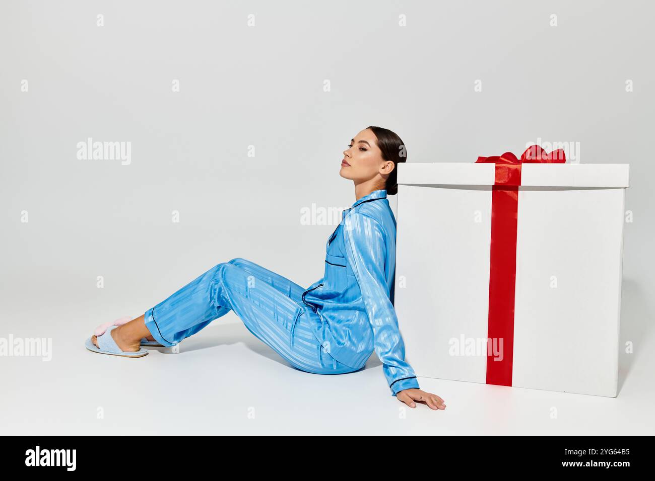 Questa bella donna si appoggia su un regalo gigante mentre indossa eleganti loungewear blu. Foto Stock