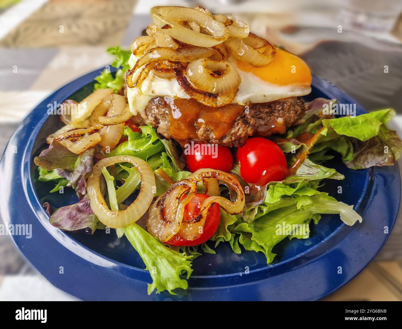 Primo piano di un piatto blu decorato ripieno di un hamburger fritto uova anelli di cipolla pomodori e lattuga con un condimento Foto Stock