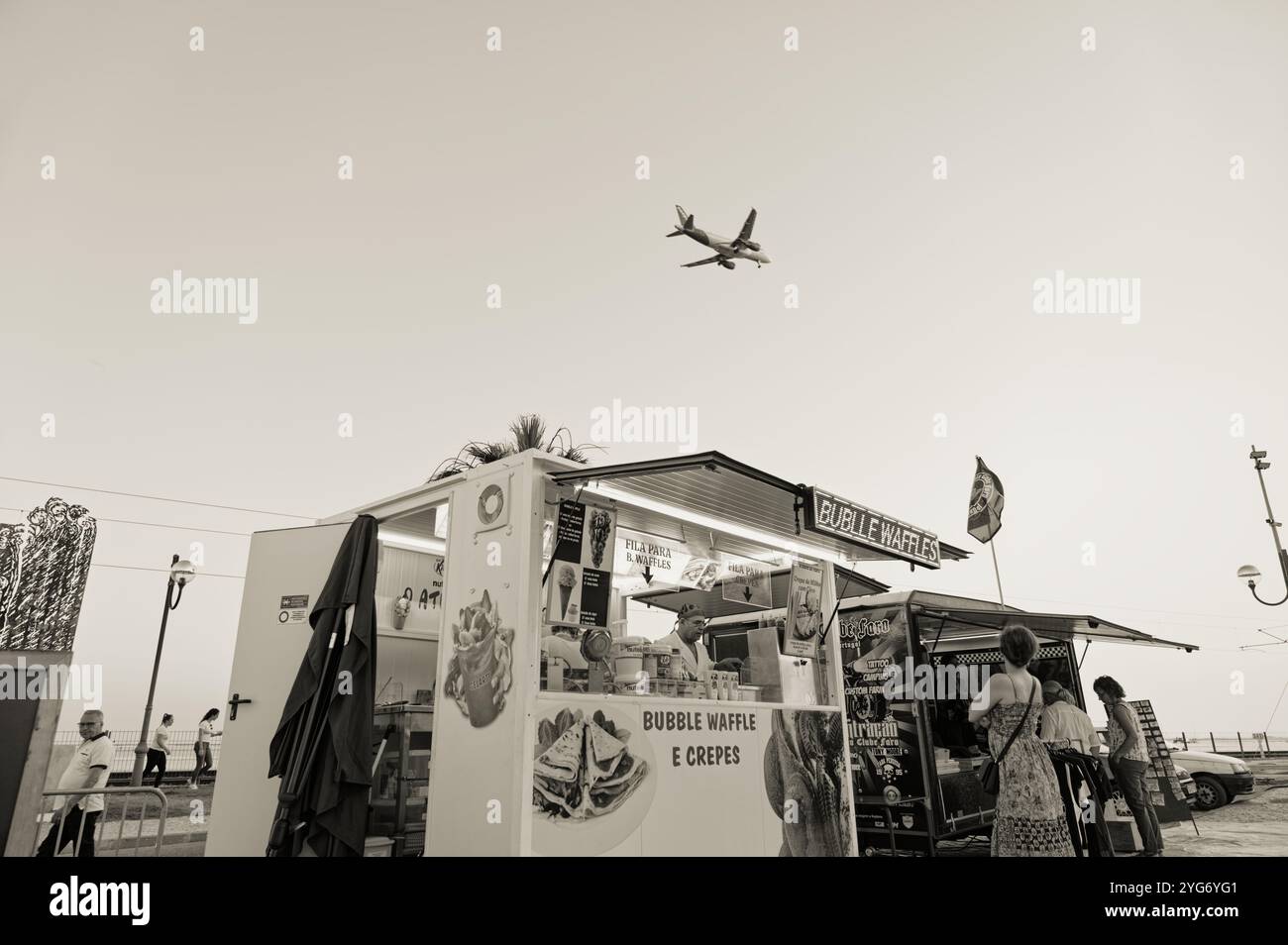 Scena monocromatica di un waffle e una crepe stall mentre un aereo vola sopra la testa in una fiera all'aperto a Faro Foto Stock