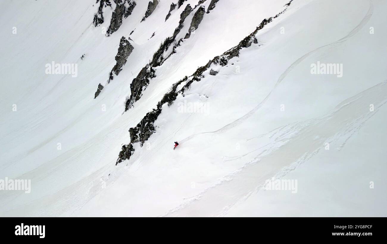 Un singolo snowboarder scivola su una ripida faccia aperta di neve pura, incorniciata da scuri affioramenti rocciosi. Foto Stock
