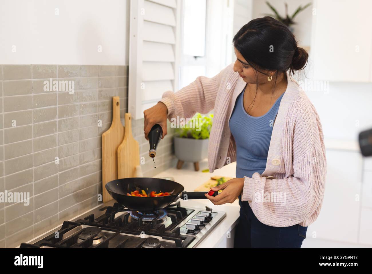 Donna asiatica che cucina verdure in cucina, aggiungendo olio per friggere in padella, a casa Foto Stock