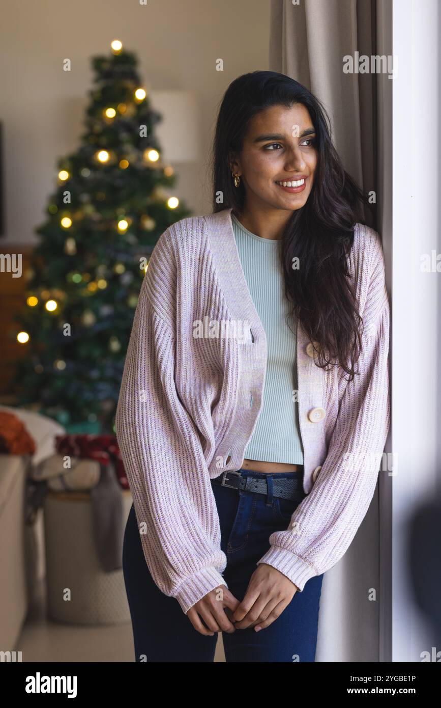 Donna asiatica sorridente in piedi accanto alla finestra, godendo delle luci festose degli alberi di Natale, a casa Foto Stock