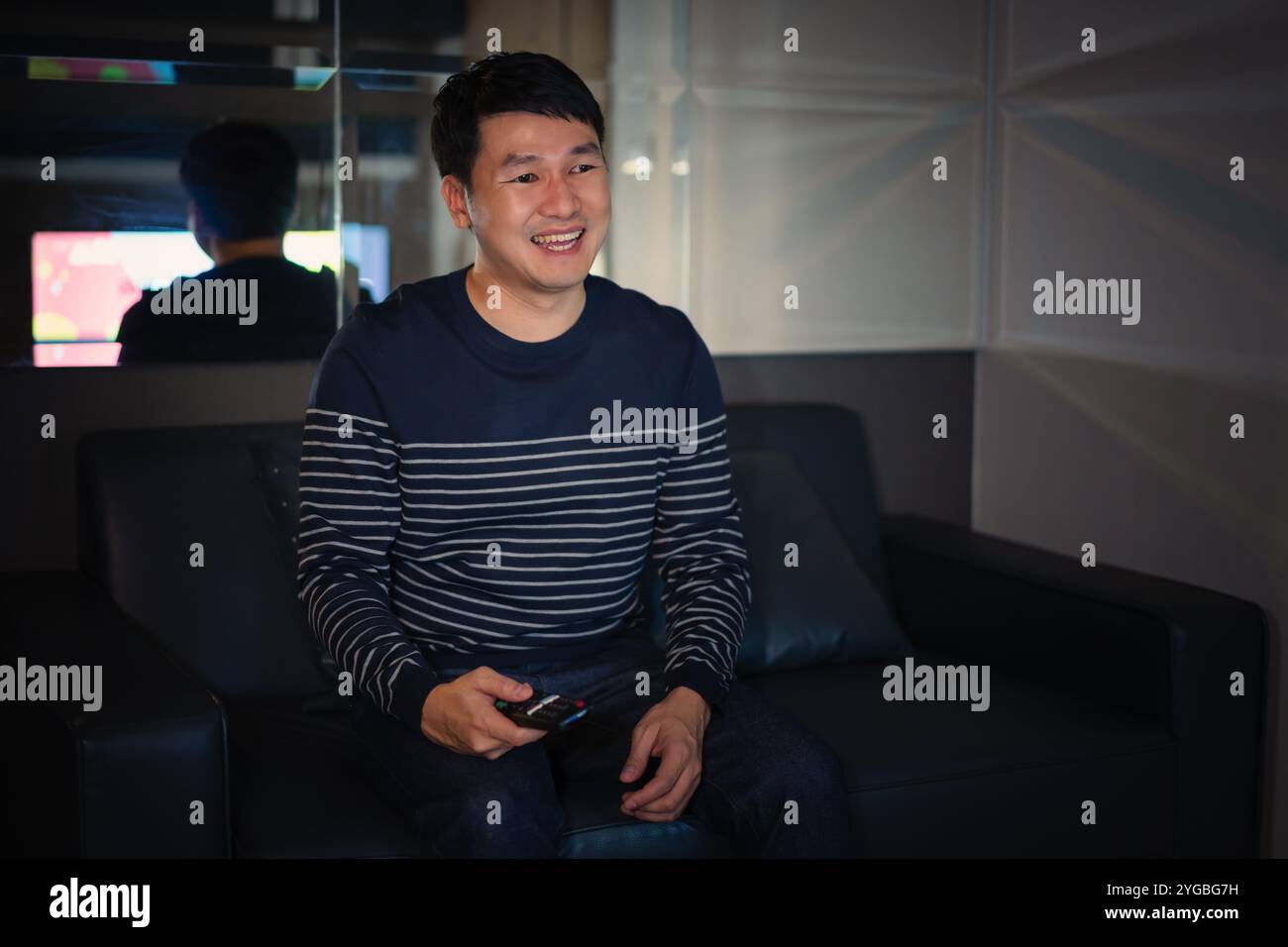 Uomo allegro che tiene in mano il telecomando e guarda la TV mentre si siede su un divano la notte Foto Stock