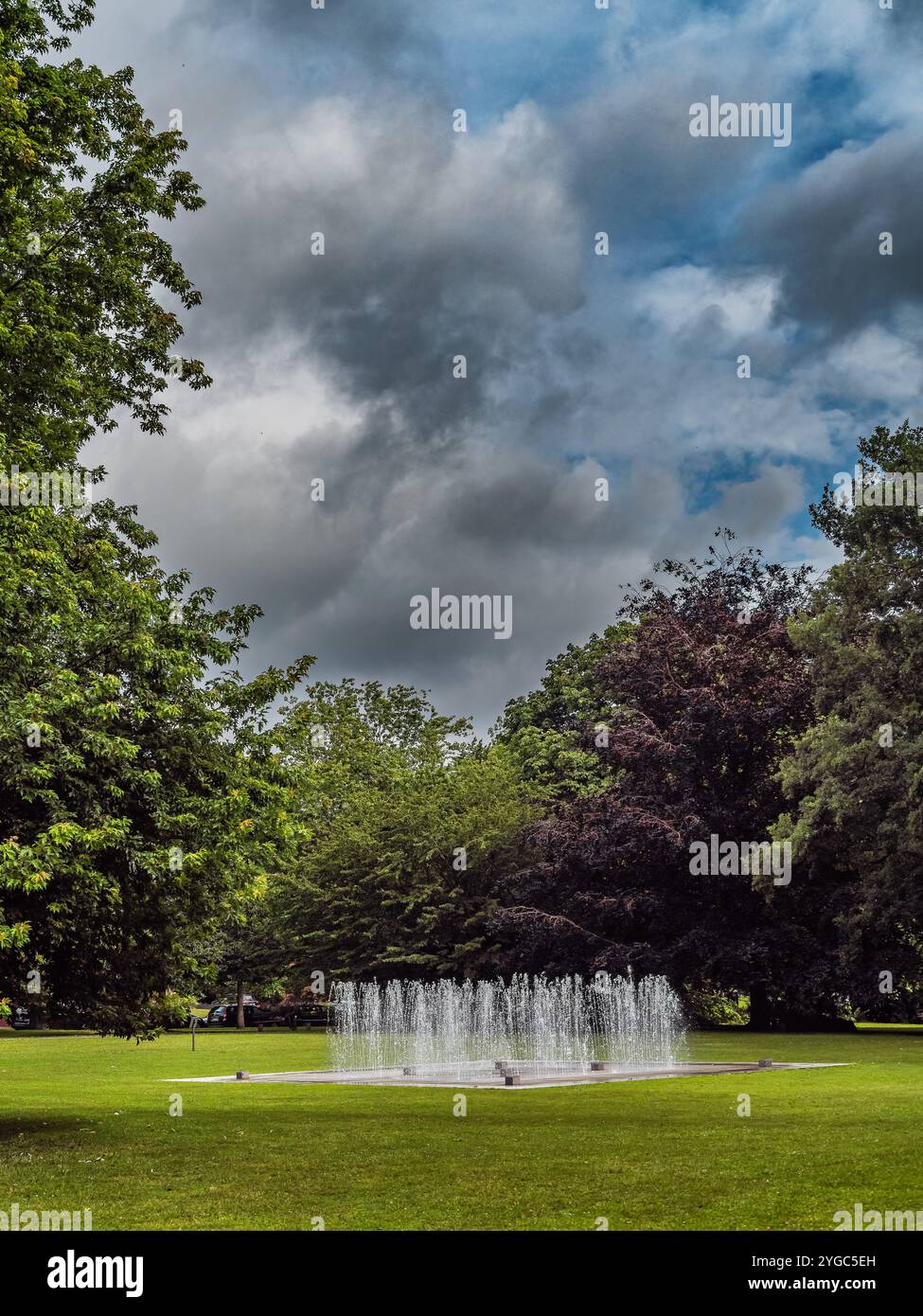 Una tranquilla fontana spruzza l'acqua in un tranquillo parco verde, circondato da alberi lussureggianti e aree erbose, offrendo un senso di relax e bellezza naturale. Foto Stock