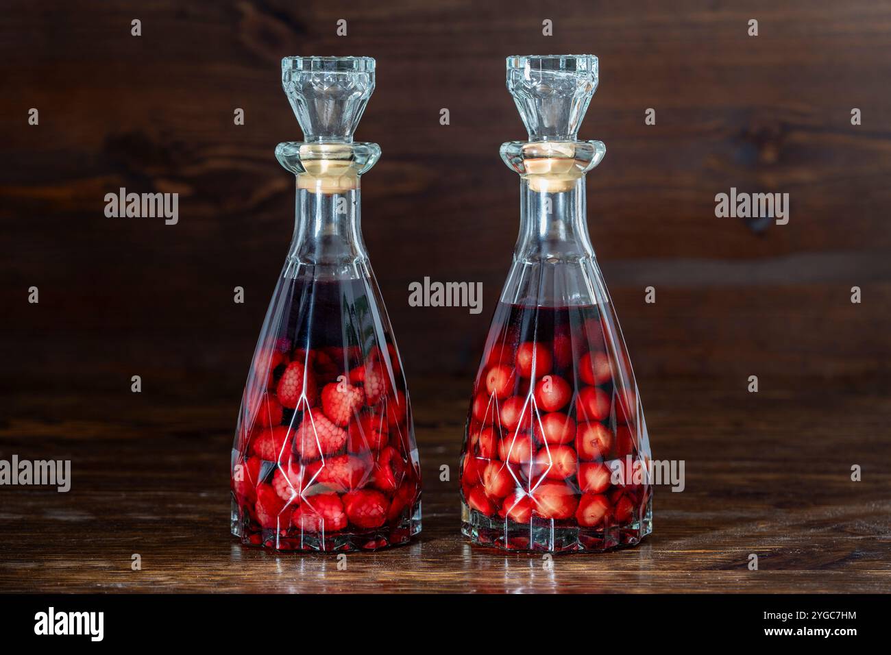 Due bottiglie di liquore di ciliegia rossa e lamponi fatti in casa su uno sfondo di legno, primo piano. Bevande alcoliche di bacca Foto Stock