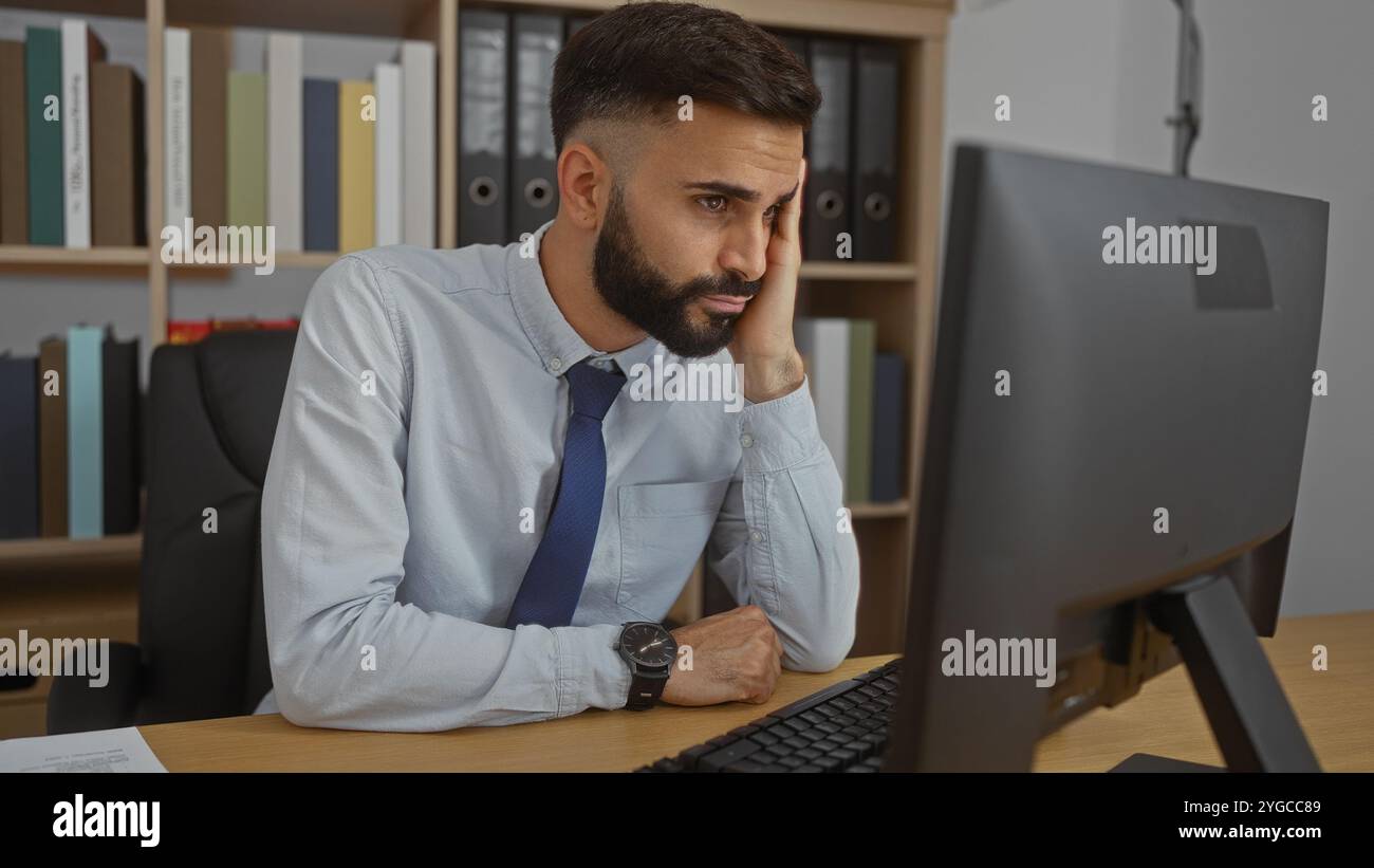 Uomo ispanico con la barba in un ufficio, seduto a una scrivania, guardando lo schermo di un computer, indossando una camicia e una cravatta, apparendo premuroso o preoccupato, wi Foto Stock