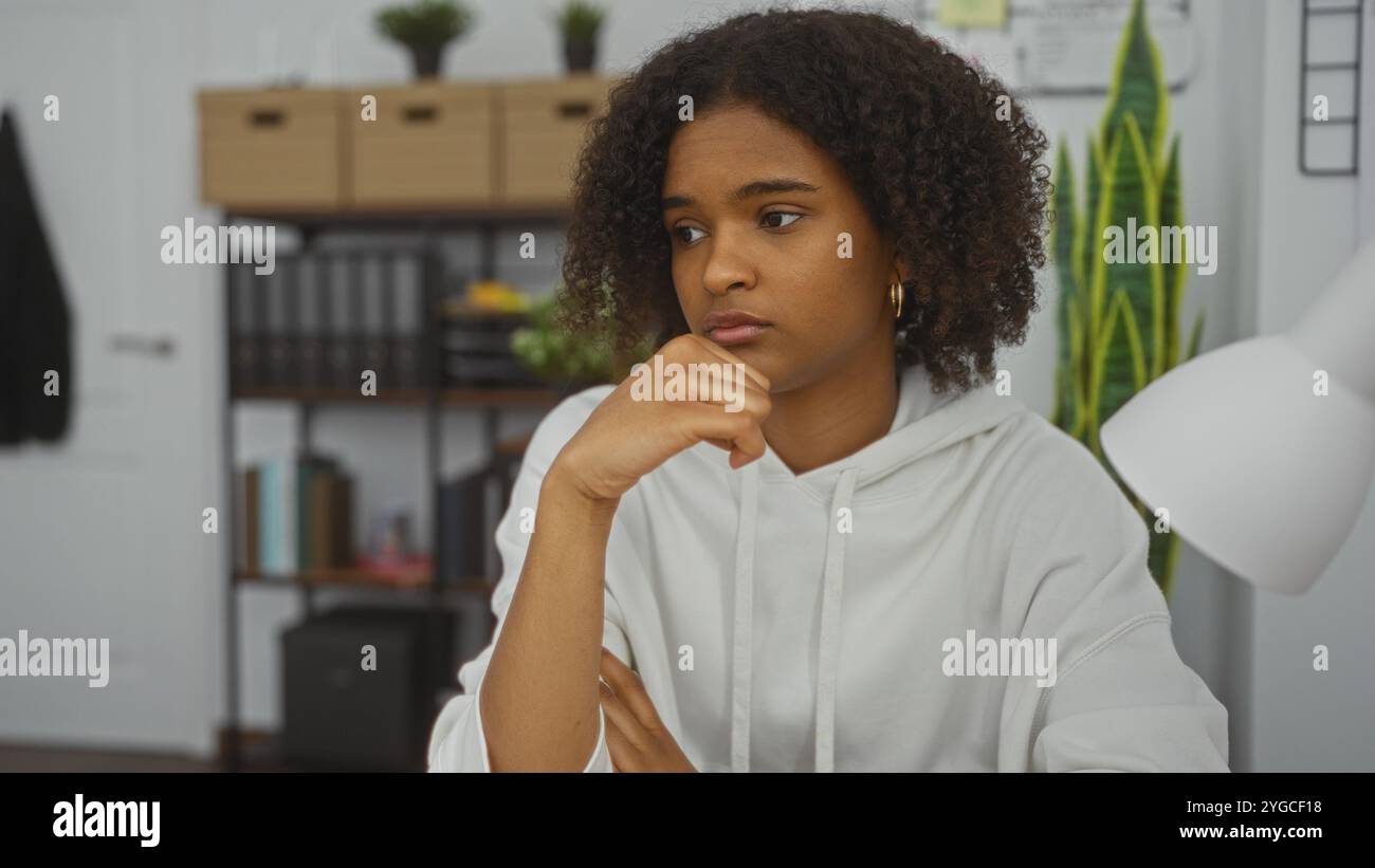 Giovane donna afro-americana con capelli ricci in un abbigliamento casual seduta con cura in un ufficio con sfondo da libreria. Foto Stock