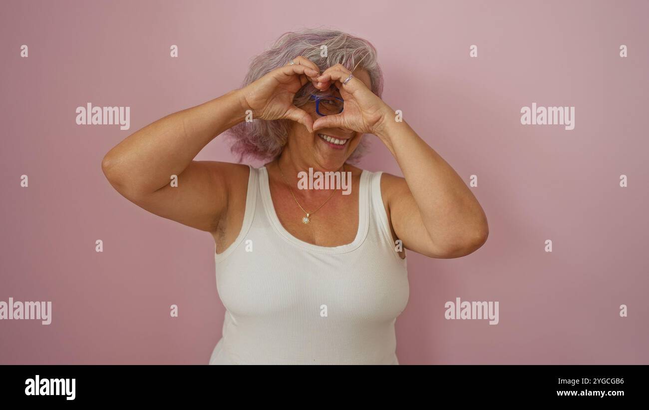 Donna anziana che forma un cuore con le mani contro un muro rosa, mostrando felicità e bellissimi capelli grigi Foto Stock