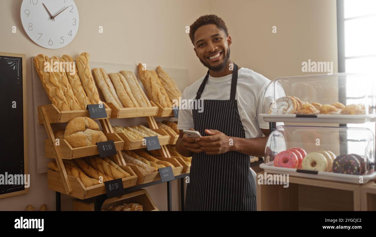 Giovane sorridente in panetteria con pane sullo sfondo e pasticcini in esposizione mentre tiene in mano un telefono Foto Stock