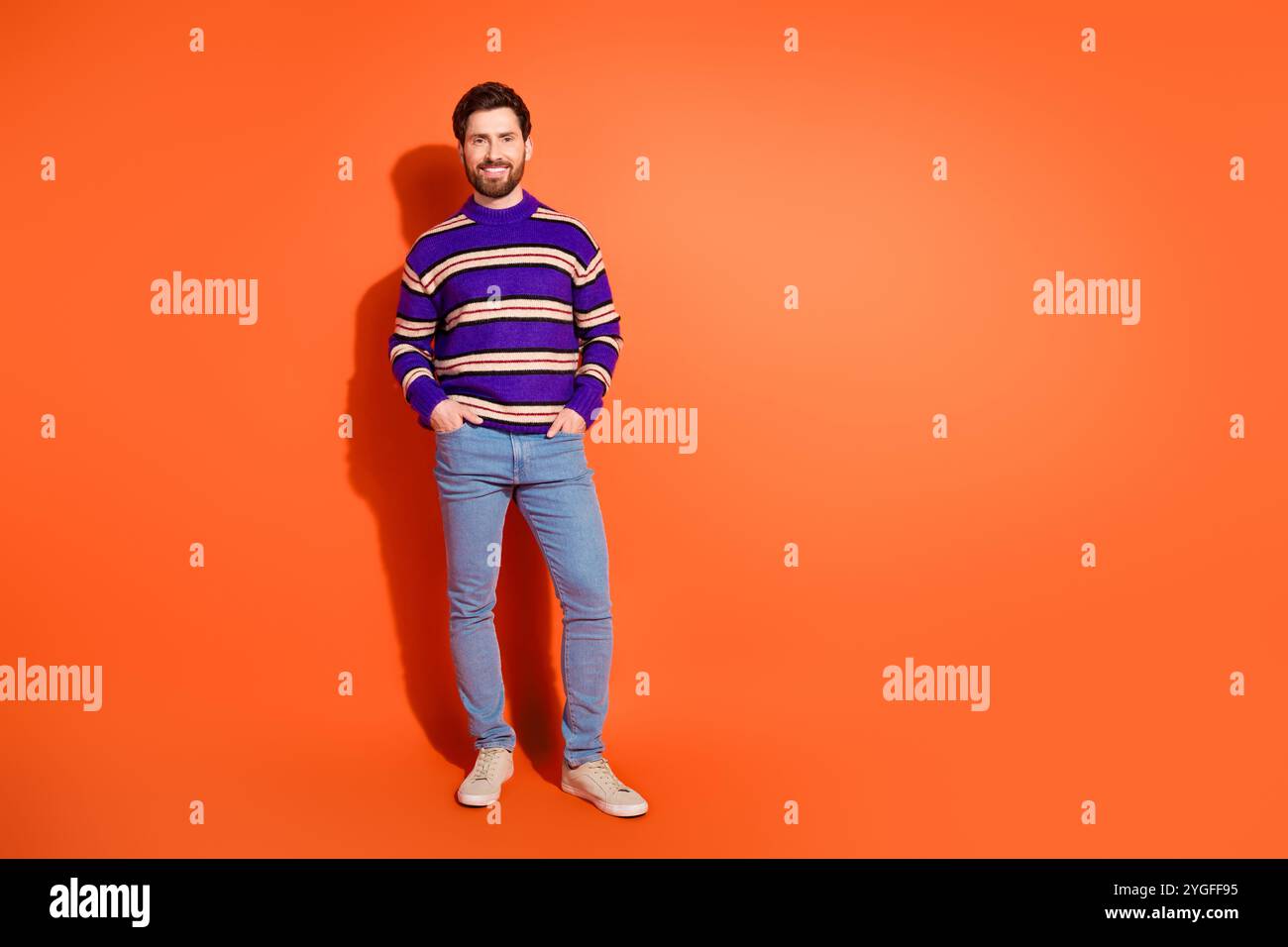 Il giovane uomo barbuto in un elegante pullover a righe si erge con sicurezza su un vivace sfondo arancione, simboleggiando la moda moderna e l'energia giovanile. Foto Stock