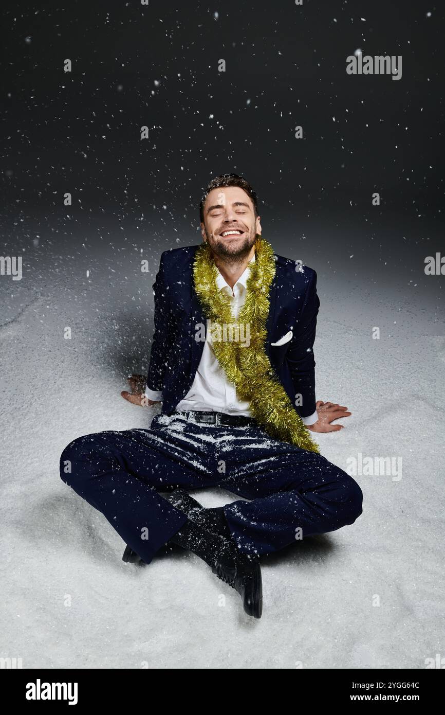 Un giovane è seduto sulla neve soffice con un sorriso allegro, adornato da decorazioni festose. Foto Stock