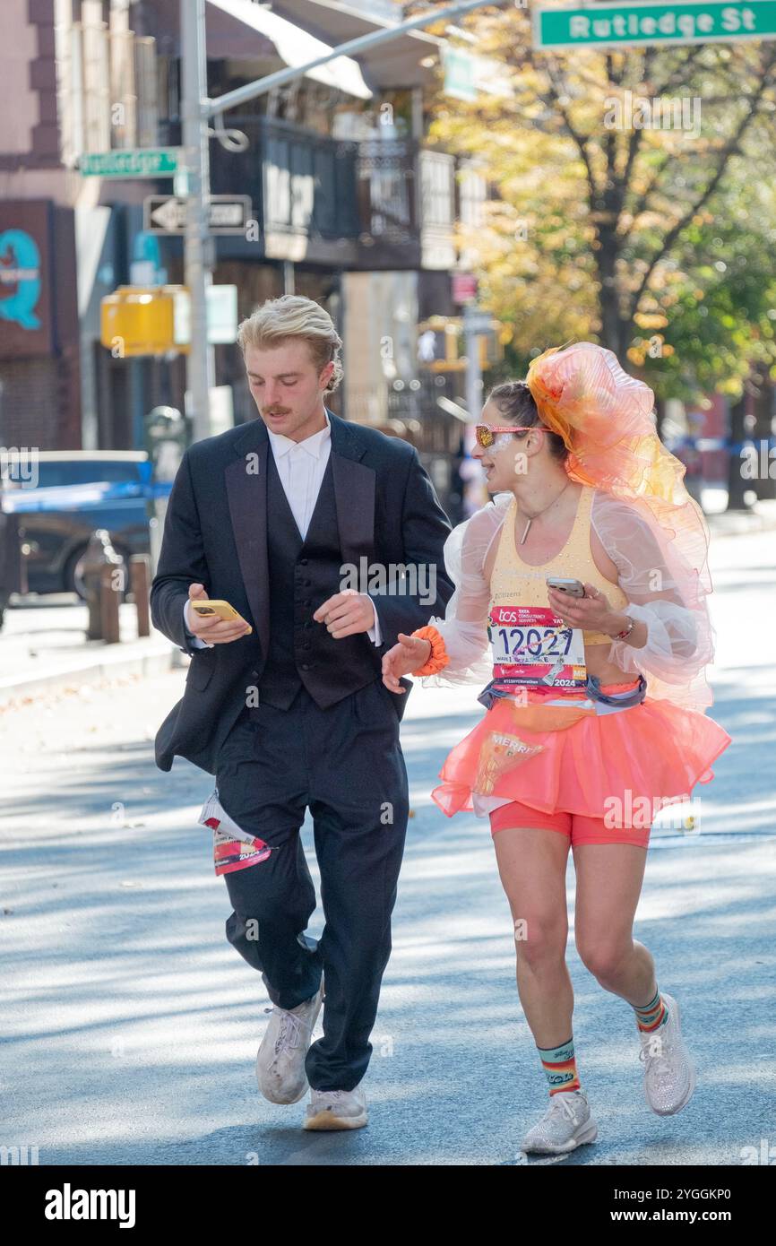 Una coppia che corre la Maratona di New York del 2024 in costumi e tiene in mano i cellulari. Su Bedford Ave a Williamsburg, circa 16 km. Foto Stock