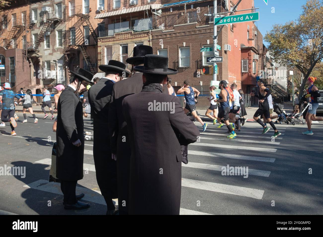Ai 16 km della maratona di New York del 2024, gli ebrei ortodossi attendono l'opportunità di attraversare Bedford Ave ammirando l'enorme corona di piloti. Foto Stock