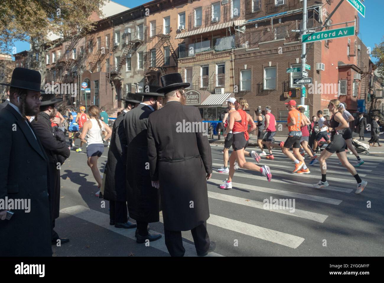 Ai 16 km della maratona di New York del 2024, gli ebrei ortodossi attendono l'opportunità di attraversare Bedford Ave ammirando l'enorme corona di piloti. Foto Stock