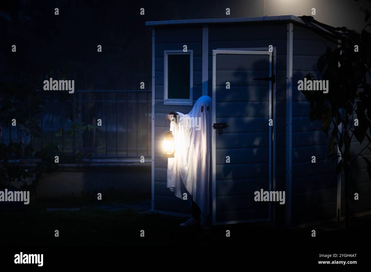 Classico costume fantasma realizzato con una coperta bianca con fori che escono da un capannone da giardino con una lampada. Spettrale sfondo di halloween con spazio per le copie. Foto Stock
