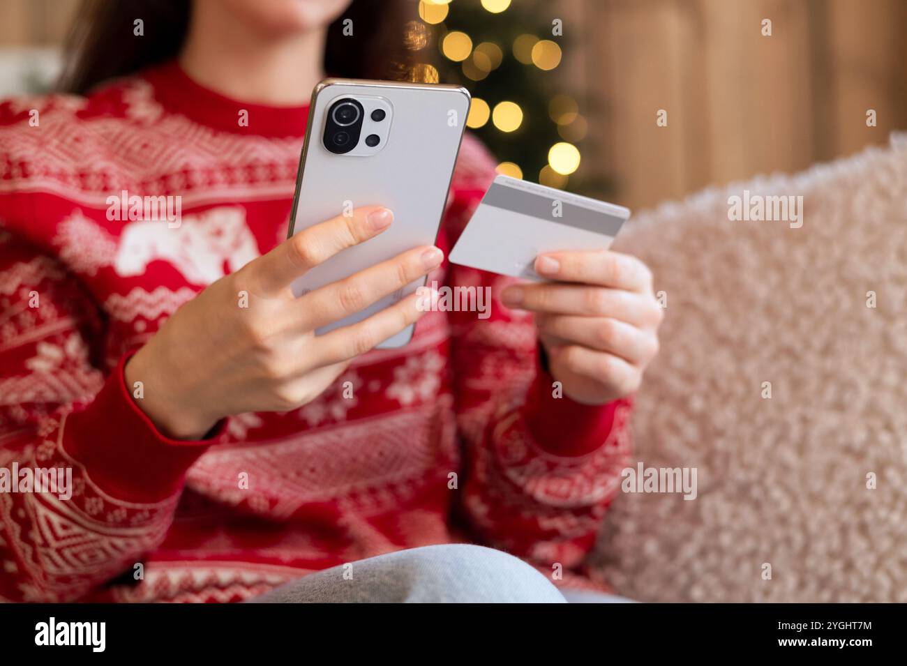 Donna in maglione natalizio che utilizza smartphone e carta di credito per lo shopping natalizio, con luci di Natale sfocate sullo sfondo. Foto Stock