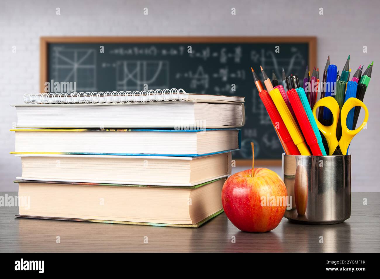 Una pila di libri di testo si trova su una scrivania accanto a una mela rossa brillante e a un contenitore pieno di cancelleria colorata, ideale per una sessione di studio in classe. Foto Stock