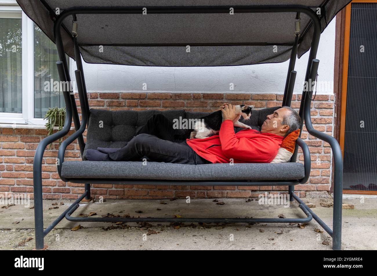 Un uomo in rosso è sdraiato su un divano oscillante nel giardino della sua casa con il suo gatto. Rilassato, confortevole, senza stress, tempo libero, non lavorare, riposante Foto Stock