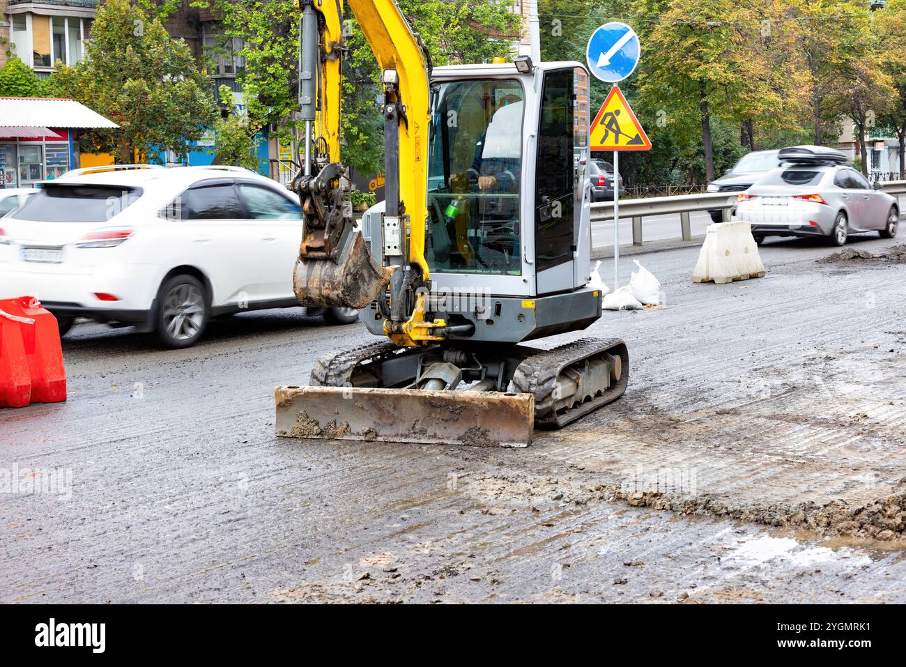 Il personale addetto alle costruzioni utilizza un escavatore compatto per riparare una strada trafficata in città in una giornata di pioggia Foto Stock