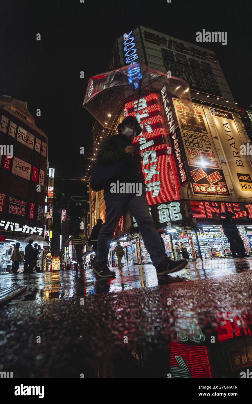 Un uomo con un ombrello passa davanti al negozio Yodobashi Kamera a Shinjuku sotto la pioggia la sera. Le luci al neon si riflettono sulla strada bagnata Foto Stock