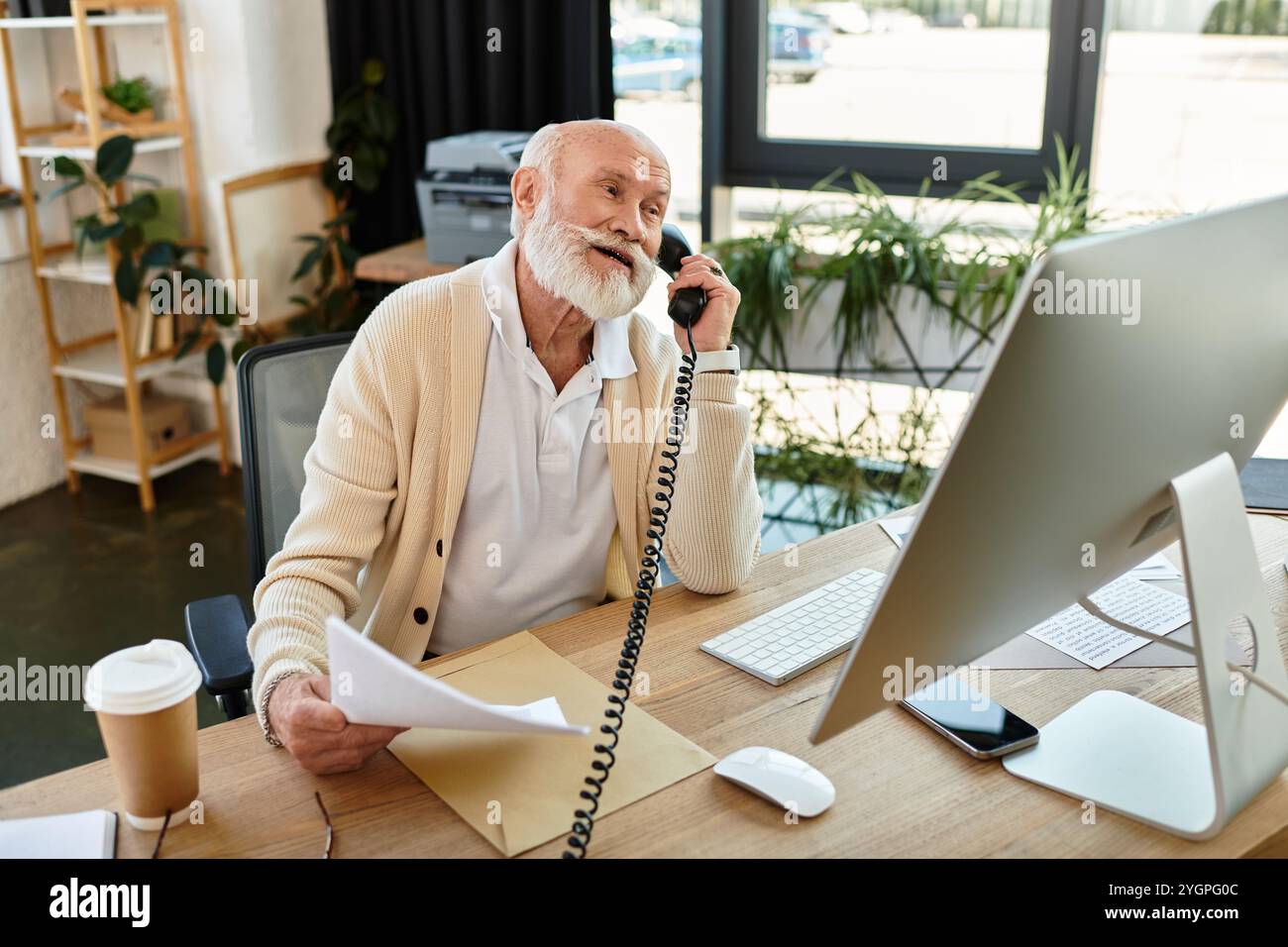 Un elegante uomo d'affari senior parla al telefono mentre si siede alla sua moderna scrivania con i documenti. Foto Stock