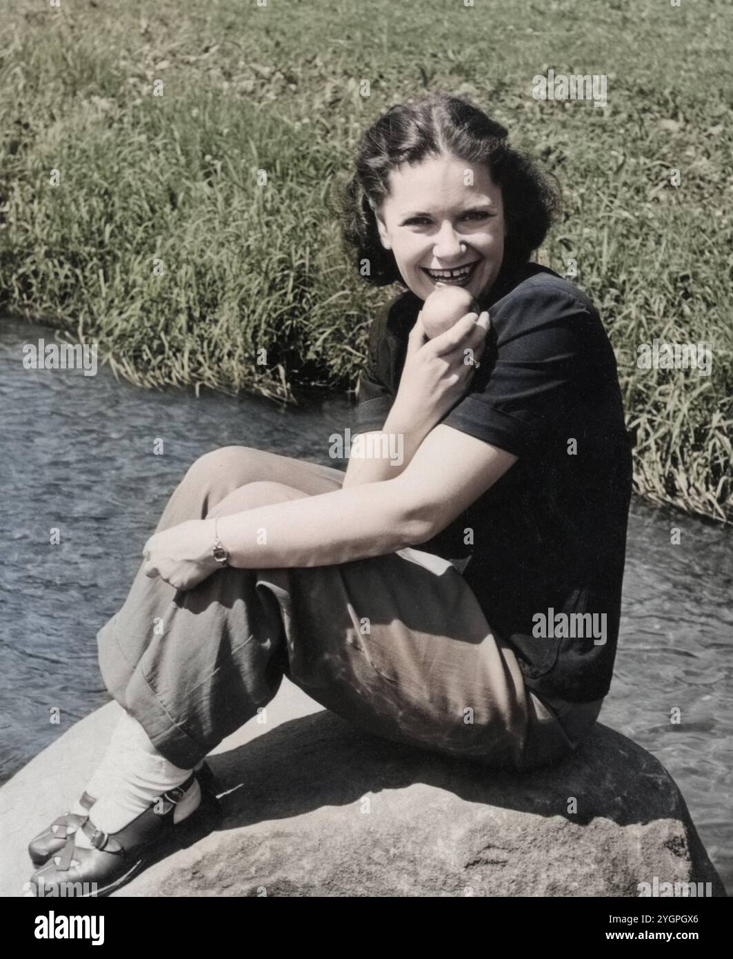 Fotografia d'epoca di una giovane donna, 27 anni, seduta su una roccia vicino a un ruscello e mangiando una mela, 1946 anni. Foto Stock