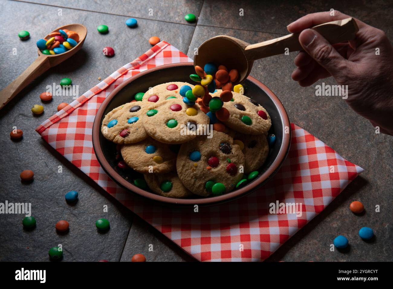 Mano dell'uomo che versa gocce di cioccolato con cucchiaio di legno sui biscotti alla vaniglia con gocce di cioccolato rosse, blu, verdi e gialle su un piatto rotondo nero su una Foto Stock