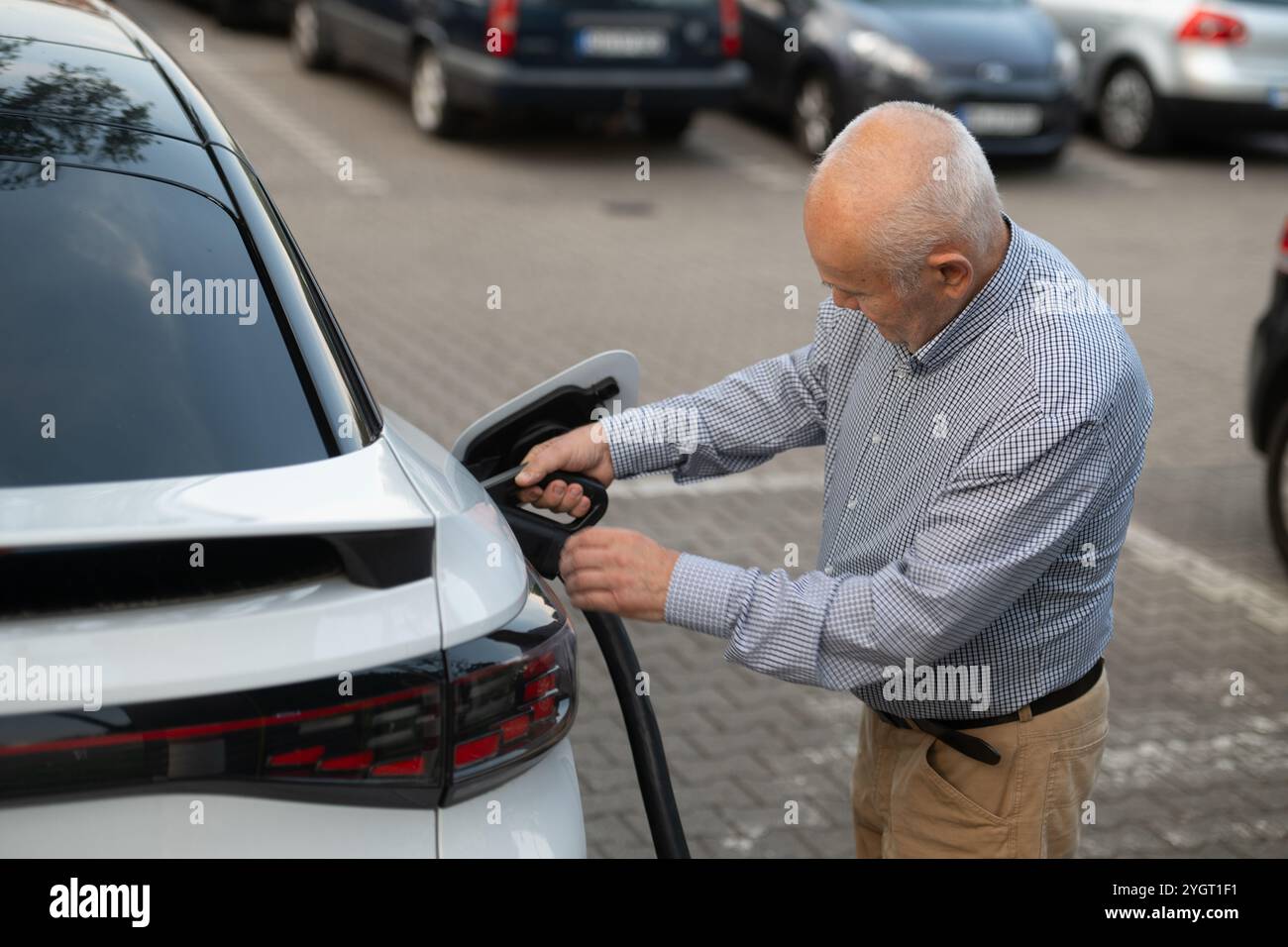 Un anziano collega un cavo di ricarica alla sua auto elettrica in un parcheggio, adottando un trasporto ecosostenibile e sostenibile. Foto Stock Un anziano collega un cavo di ricarica alla sua auto elettrica in un parcheggio, adottando un trasporto ecosostenibile e sostenibile. Foto Stock