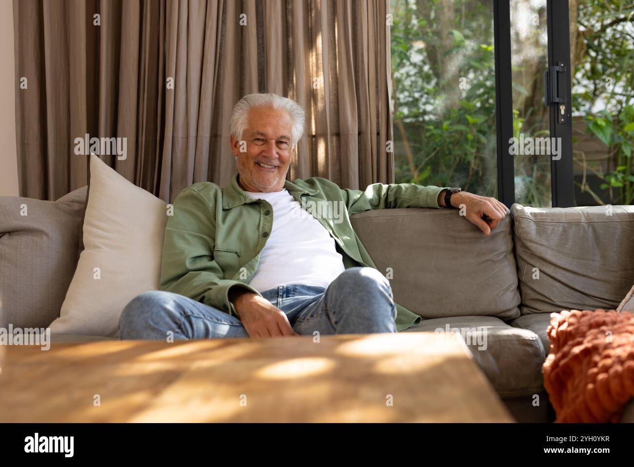 Un anziano sorridente che si rilassa sul divano a casa, godendosi il sole del pomeriggio Foto Stock