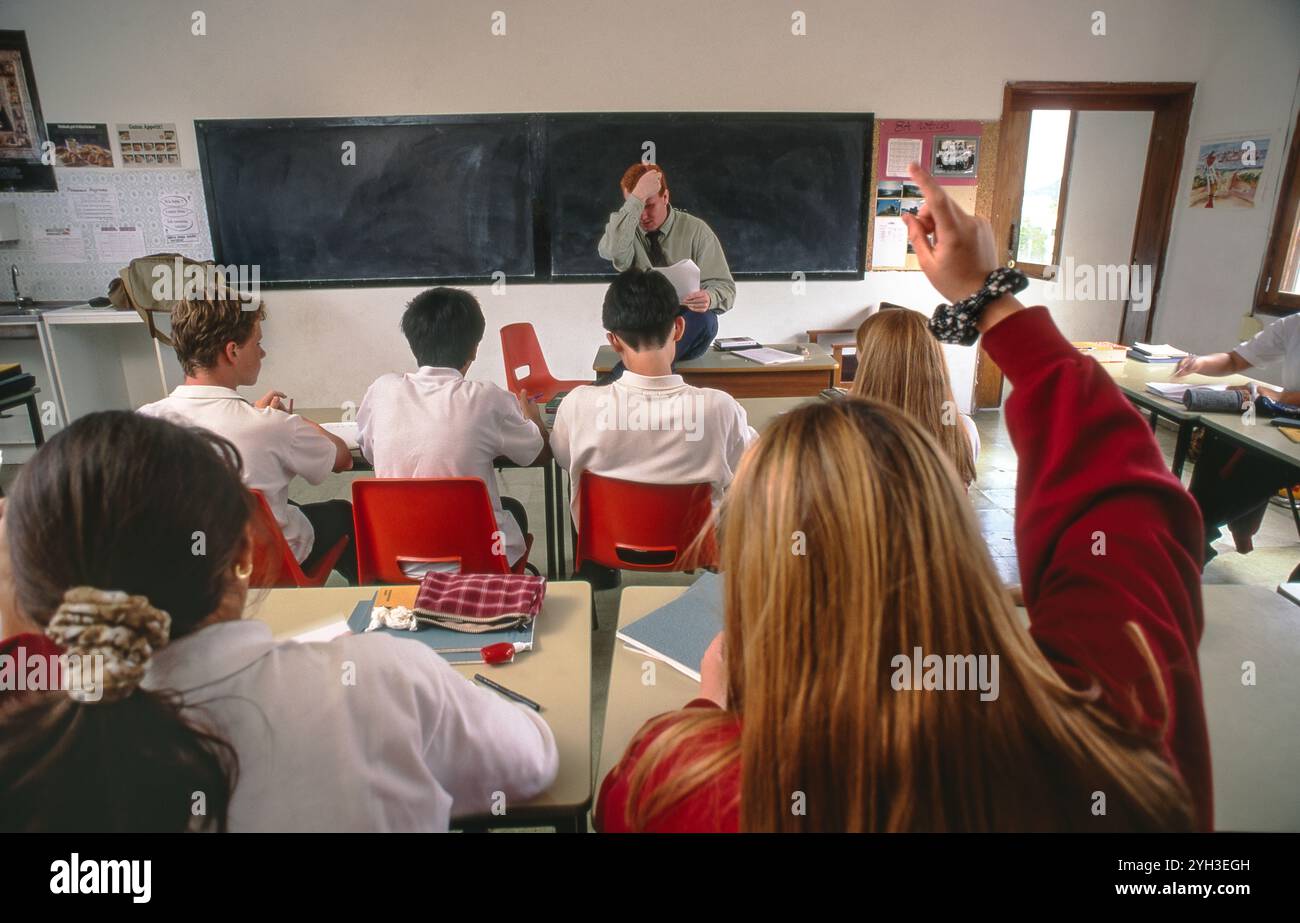 Studenti anziani di 15-17anni in classe con insegnante di sesso maschile incerto con carta davanti alla classe con lavagna. Studentessa adolescente con la mano alzata in primo piano. Foto Stock