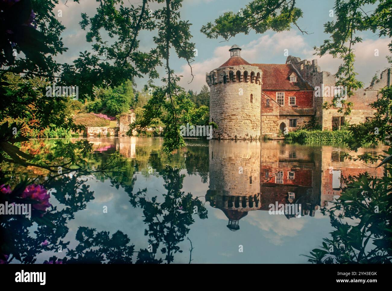 Scotney Castle rovine mistiche d'epoca retrò, viste attraverso rododendri con riflessi sull'acqua ferma sul fossato del castello, Lamberhurst, Kent, Regno Unito Scotney Castle è una casa di campagna inglese con giardini formali a sud-est di Lamberhurst nella valle del fiume Bewl nel Kent, in Inghilterra. I giardini, che sono un sito di particolare interesse scientifico e un famoso esempio di stile pittoresco, sono aperti al pubblico. Foto Stock