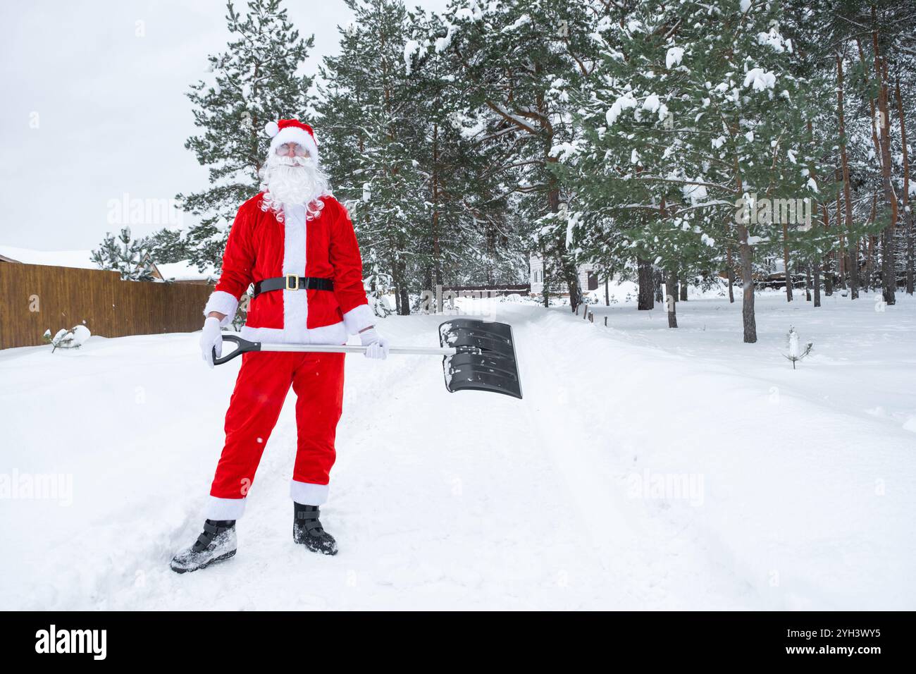 Babbo Natale pulisce la neve con la pala in inverno all'aperto dopo una nevicata. Pulire le strade del villaggio, pulire il passaggio per le auto, difficile Foto Stock