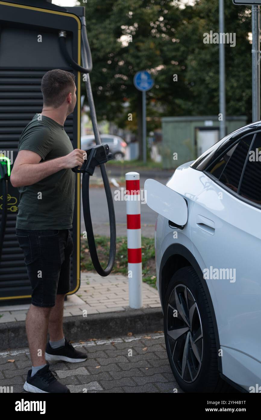 Un uomo ha in mano un connettore di carica EV, preparandolo a collegarlo a un'auto elettrica bianca in una stazione di ricarica pubblica. Foto Stock Un uomo ha in mano un connettore di carica EV, preparandolo a collegarlo a un'auto elettrica bianca in una stazione di ricarica pubblica. Foto Stock