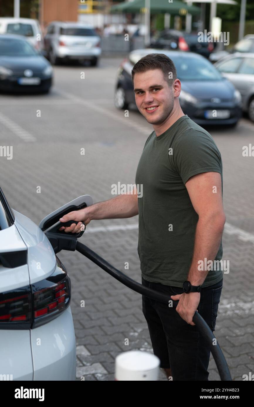 Un uomo sorridente carica un'auto elettrica in una stazione di ricarica in un parcheggio pubblico. Foto Stock Un uomo sorridente carica un'auto elettrica in una stazione di ricarica in un parcheggio pubblico. Foto Stock