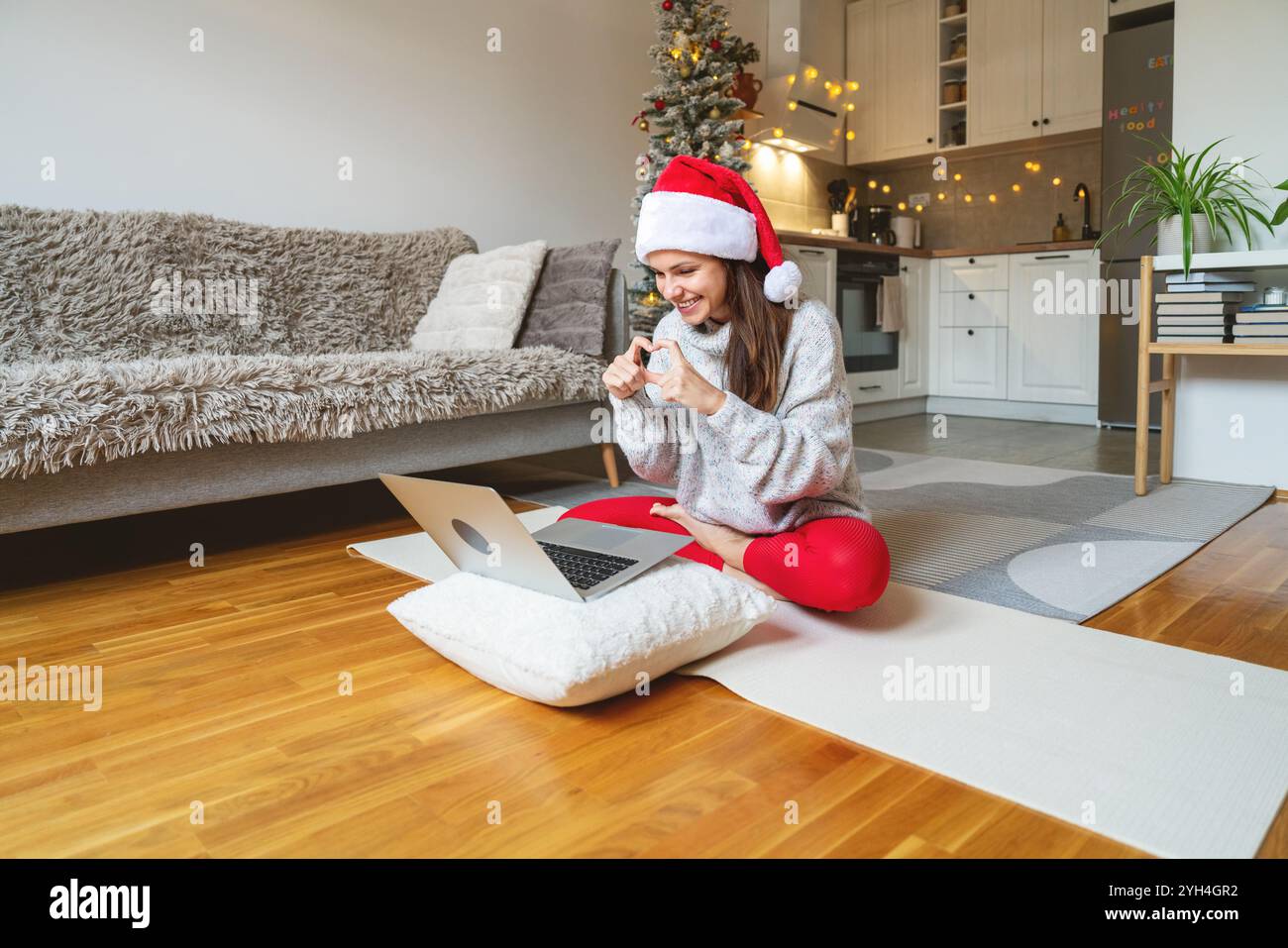 Una donna con il cappello di Babbo Natale siede sul pavimento in posa di loto, usando il laptop per videochiamate e gesti con le mani del cuore. Foto Stock