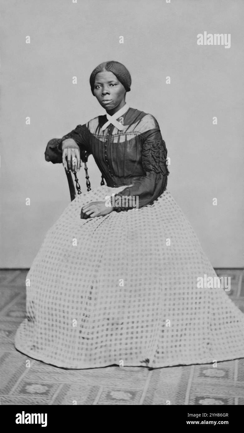 Una carta-de-visite di Harriet Tubman (circa 46-47) nel 1868-69. E' seduta, girata verso sinistra. Una mano poggia sul retro di una sedia di legno, un'altra sulle sue ginocchia. Creato da Benjamin Powelson, 77 Genesee St., Auburn, New York. Harriet Tubman seduto in una stanza interna. È posizionata leggermente girata a destra e guarda fuori dalla telecamera. La sua mano destra poggia sul retro di una sedia di legno e la sua mano sinistra è in grembo. Un tappeto dai motivi audaci copre il pavimento e la parete o la goccia dietro di lei è di colore chiaro vuoto. Tubman indossa un corpetto nero che si abbotta nella parte anteriore centrale e. Foto Stock