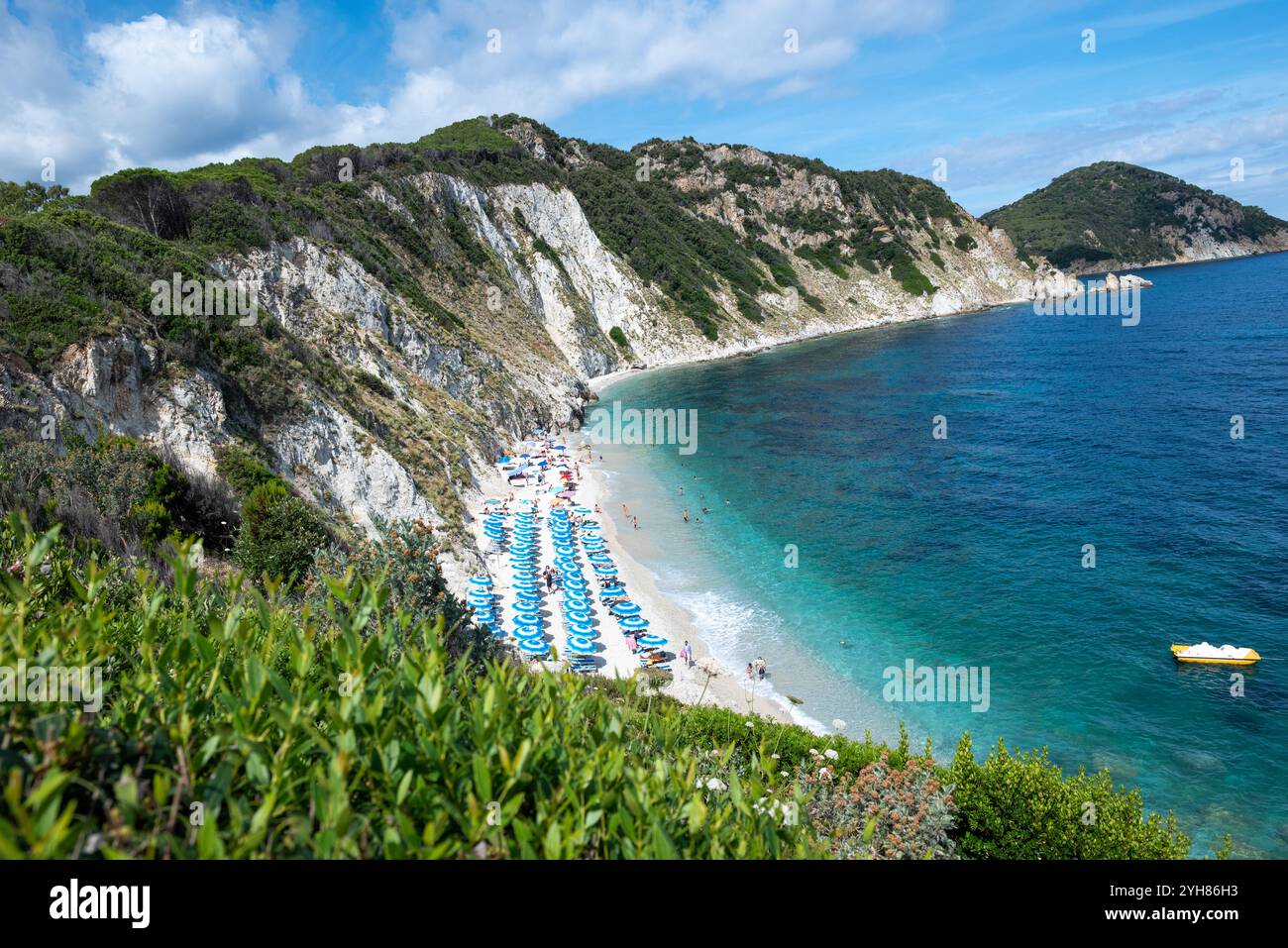 Spiaggia di Sansone all'Isola d'Elba in estate, con lettini e ombrellone con molti bagnanti in riva al mare e pedalo giallo all'ancora Foto Stock