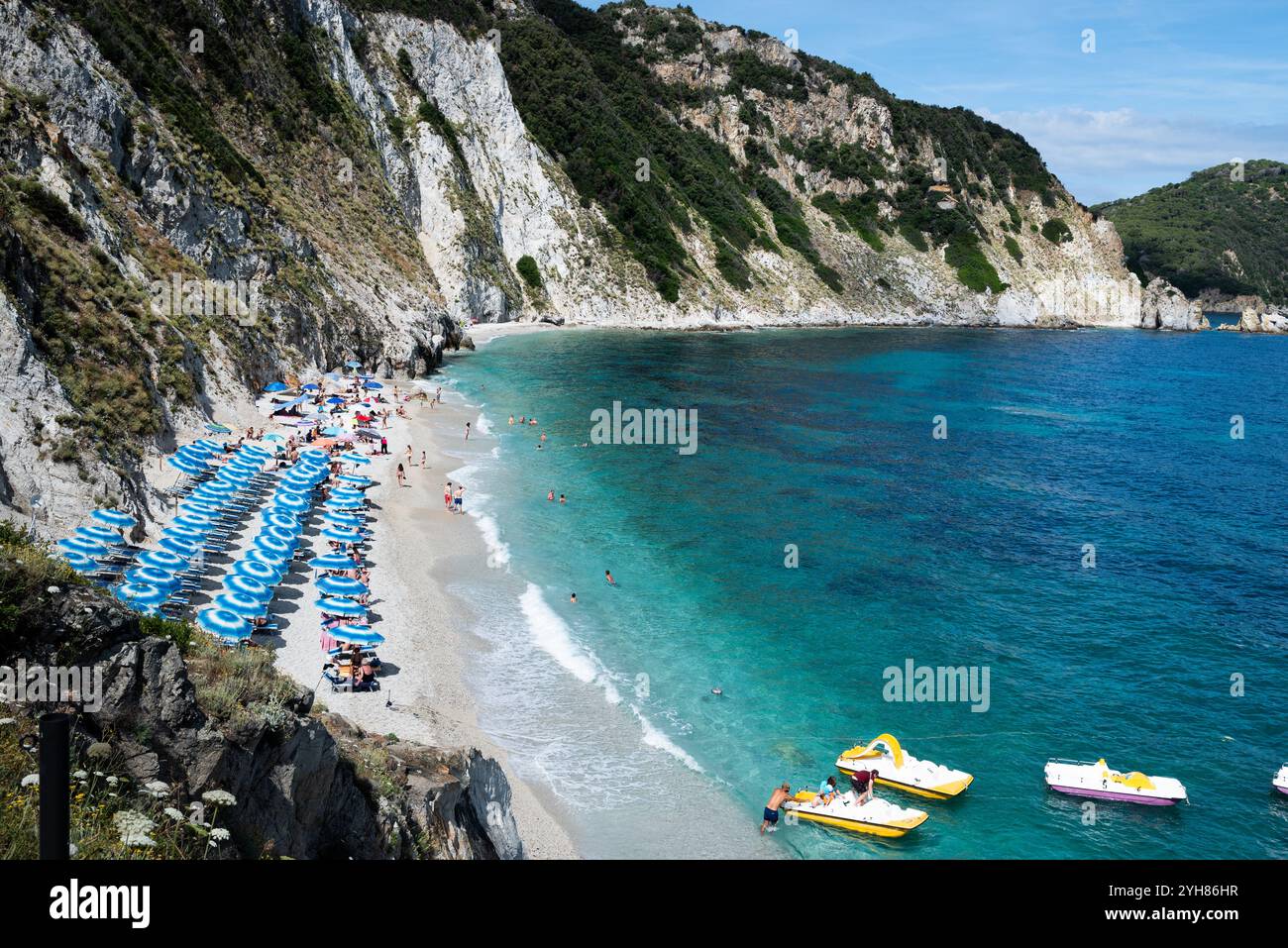 Spiaggia di Sansone all'Isola d'Elba in estate, con lettini e ombrellone con molti bagnanti in riva al mare e barche a remi colorate Foto Stock