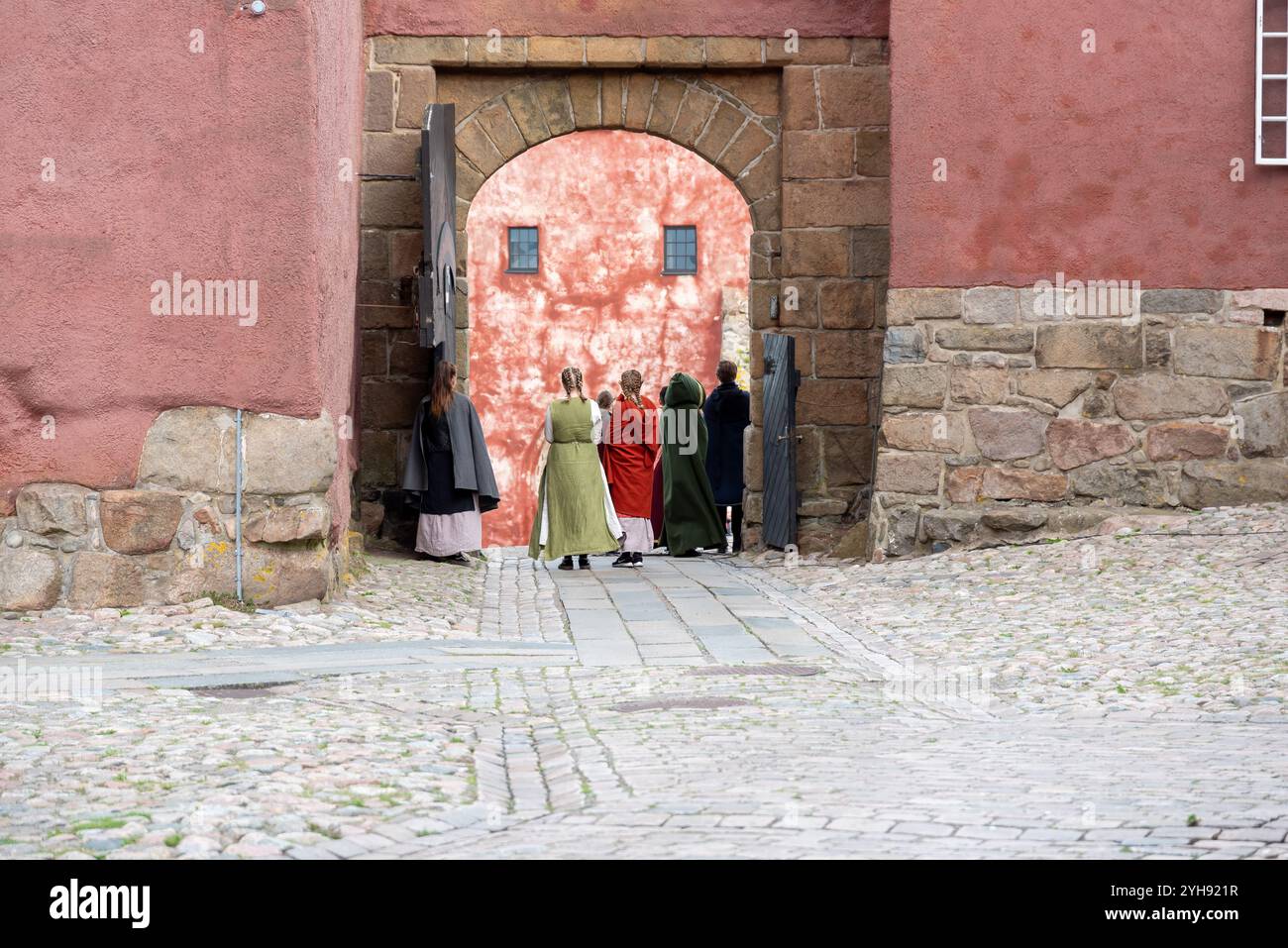 Un gruppo di individui vestiti con costumi storici cammina verso un'imponente porta rossa di legno di un castello medievale. Il percorso acciottolato si aggiunge al Foto Stock