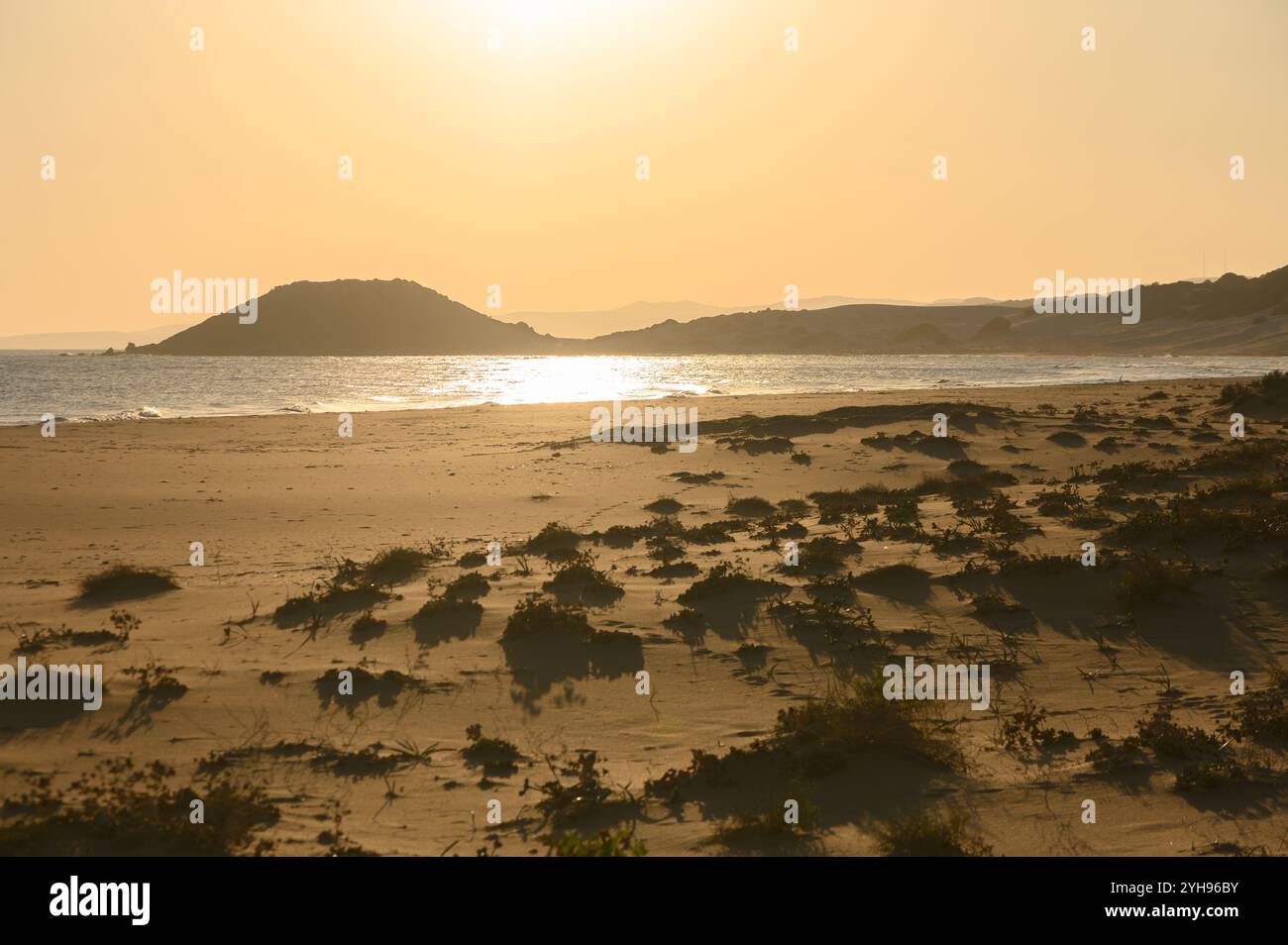 Il sole tramonta su una spiaggia tranquilla, proiettando ombre morbide sulla spiaggia sabbiosa e illuminando colline lontane. Foto Stock