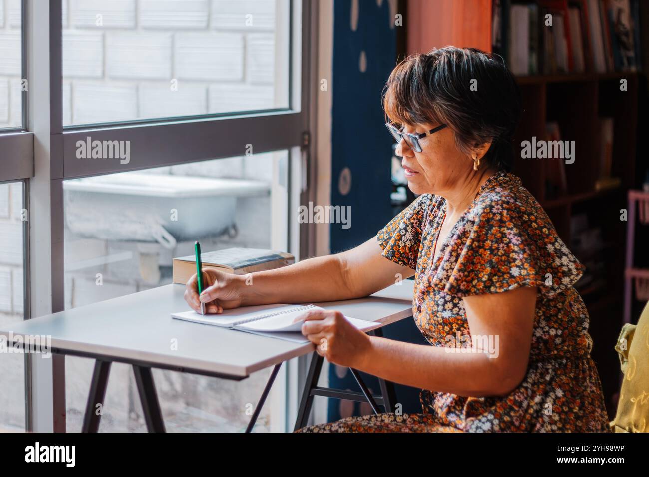 Una donna matura con i capelli corti, vestita con un abito floreale, seduta a una scrivania e scrivendo con attenzione nel suo notebook, godendosi un momento di tranquillità. Foto Stock